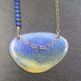 This ia a Pendant Necklace with an enamelled blue and yellow surface with a dark blue speckled effect. It is an irregular elipical shape. The colours intermingle with lavender blue at the top of the pendant and sandy yellow at the bottom. A darker blue creates a speckled effect from top to bottom of the pendant. The pendant hangs on a gold chain on one side and a string of small round lapid lazuli beads on the other. Only the pendant and a few of the beads and a portion of the  chain is visible.