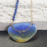 This ia a Pendant with an enamelled blue and yellow surface with a dark blue speckled effect ontop. It is an irregular elipical shape. The colours intermingle with lavender blue at the top of the pendant and sandy yellow at the bottom. A darker blue creates a speckled effect from top to bottom of the pendant. The pendant hangs on a gold chain on one side and a string of small round lapid lazuli beads on the other. Only the pendant and a few of the beads and a portion of the  chain is visible.