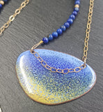 Pendant with an enamelled blue and yellow surface with a dark blue speckled effect ontop. It is an irregular elipical shape. The colours intermingle with lavender blue at the top of the pendant and sandy yellow at the bottom. A darker blue creates a speckled effect from top to bottom of the pendant. The pendant hangs on a gold chain on one side that extends over the pendant to the other side and a string of small round lapid lazuli beads on the other interspersed with gold beads.