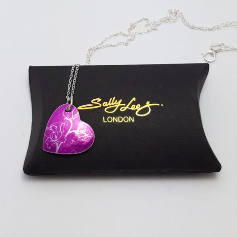 Pink heart pendant with silver coloured linear print of sweet pea flowers on a pink back ground with a silver chain. The pendant is on a pillow shaped black gift box with the Sally Lees logo in the centre in gold.
