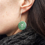 This photograph shows the green side of the reversible women's suffrage earrings. These earrings are circular with the pattern of the end of a scroll in a silvery colour on a green background. They hang on a silver hoop and are photographed on a model.