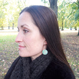 Photograph of the full head and shoulders of a model wearing these reversible circular drop earrings with sterling silver hoop wires. The side of the circular earrings are green with a silvery pattern of a scroll on them.
