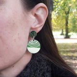 Sufragette drop stud earrings. Earrings are a small disk with  a large disk connected by a silver jump ring. The pattern is on the top of the small disk and covers half of the larger lower disk. Worn by a model.