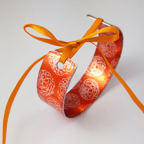 Bright orange cuff with marigold flower illustrations in a silvery colour reapeted on the inside and outside of the cuff. The ends of the cuff are visible with a hole in each end centrally placed and an orange ribbon through the holes and tied in a bow 