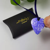 Back of a purple Guitar pick pendant decorated with a delicate linear print in a silvery colour of larkspur flowers. The pendant has a purple satin ribbon and is held in a human finger and thumb above a black pillow shaped box with the Sally Lees (London) logo in gold printed onto the box.