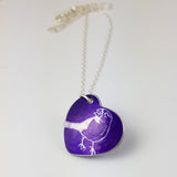 Purple heart shaped pendant with a line drawing of a cute finch seen from the side printed in a silvery colour covering the surface of the heart. The bird has a berry in its beak. The pendant is on a silver chain.