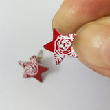 Aluminium star stud earrings with red background and abstracted rose pattern in a silvery colour. One stud is held between finger and thumb.