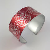 Side view of an aluminium cuff with large scroll motifs in a row over the surface in a silvery colour with  red background. The cuff is silvery coloured on the inside.