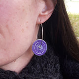 This photogrph shows the purple side of these reversible Women's suffrage circular drop hoop earrings that are patterned with the silvery pattern of a scroll hanging from a sterling silver hoop and is photgraphed on a model.