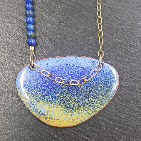 This ia a Pendant Necklace with an enamelled blue and yellow surface with a dark blue speckled effect. It is an irregular elipical shape. The colours intermingle with lavender blue at the top of the pendant and sandy yellow at the bottom. A darker blue creates a speckled effect from top to bottom of the pendant. The pendant hangs on a gold chain on one side and a string of small round lapid lazuli beads on the other. Only the pendant and a few of the beads and a portion of the  chain is visible.