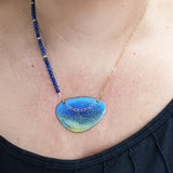 This ia a Pendant with an enamelled blue and yellow surface with a dark blue speckled effect ontop. It is an irregular elipical shape. The pendant hangs on a gold chain on one side and a string of small round lapid lazuli beads and gold beads on the other. The chain also hangs over the front of the pendant. The pendant is photographed on a human necklace.