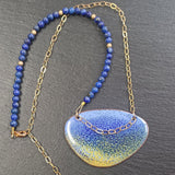 Pendant with an enamelled blue and yellow surface with a dark blue speckled effect ontop. It is an irregular elipical shape. The colours intermingle with lavender blue at the top of the pendant and sandy yellow at the bottom. A darker blue creates a speckled effect from top to bottom of the pendant. The pendant hangs on a gold chain on one side that extends over the pendant to the other side and a string of small round lapid lazuli beads on the other interspersed with gold beads. 