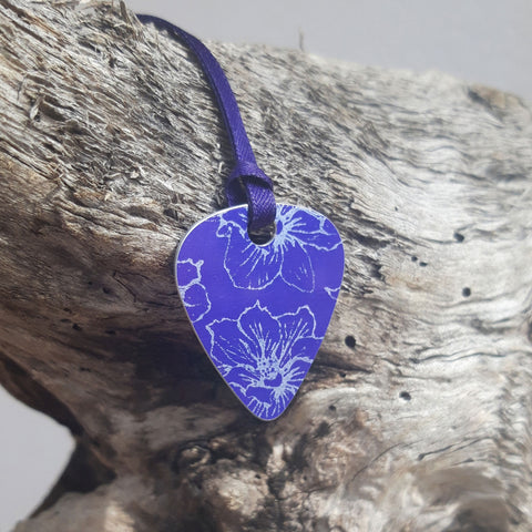 Front view of a purple Guitar pick pendant decorated with a delicate linear print in a silvery colour of larkspur flowers. The pendant has a purple satin ribbon and is hung over a wooden log that has been bleached by the sea. 