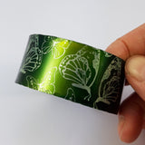 Anodized aluminum cuff printed with butterflies and dyed a dark green and lighter green. This photograph shos the lighter green side held in the artist's hand.