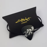 Adjustable anodised aluminium guitar pick shaped bracelet with faux leather cord. The guitar pick is black with silvery musical notes pattern across the middle of the pick.