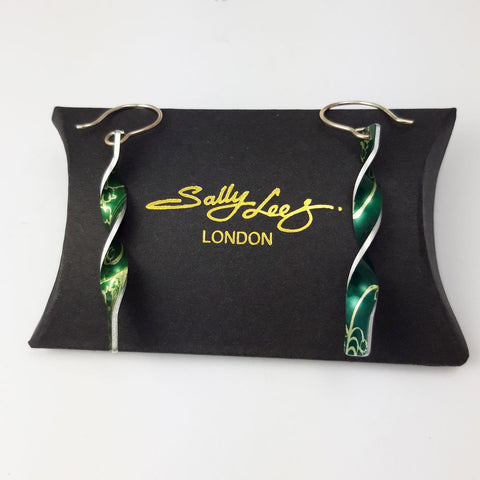 Green aluminium twist earrings with a lily of the valley flower on both sides with silvery coloured edges displayed on a black pillow shaped box with a gold embossed Sally Lees London logo on it.