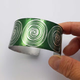 Anodized aluminum digitally printed cuff with large motifs of the end of scrolls in a linear pattern of concentric circles in a silvery colour on a green background. The inside of the cuff is a silver colour.