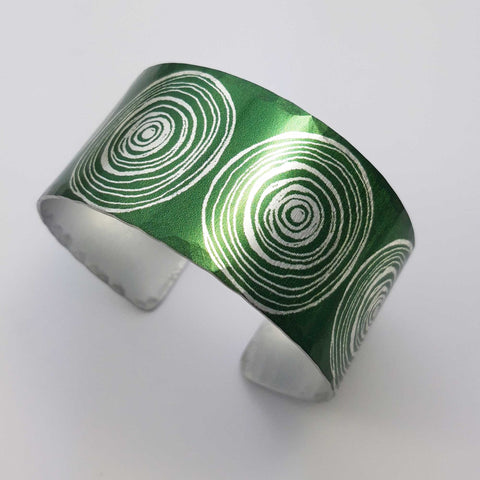 Anodized aluminum digitally printed cuff with large motifs of the end of scrolls in a linear pattern of concentric circles in a silvery colour on a green background. The inside of the cuff is a silver colour.