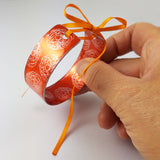 Bright orange cuff held in a hand with marigold flower illustrations in a silvery colour reapeted on the inside and outside of the cuff. The ends of the cuff are visible with a hole in each end centrally placed and an orange ribbon through the holes and tied in a bow 