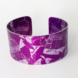 Front view of a cuff with dark magenta background with swallows butterflies and delicate bird cages illustrations in a silvery colour. The butterflies overlap the bird cages and the birds appear to fly up from the bottom of the cuff to the top of the cuff. The patterned inside of the cuff can also be viewed on this photograph.