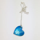 anodised aluminium heart shaped pendant with silvery delicate print of a lily of the valley flower and leaves on a dyed blue back ground with a silver chain
