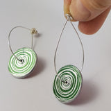 Round anodised aluminium womens suffrage earrings in green with  circular silvery coloured scroll motif on each disk. The ear wires are long and eliptical shaped and thread through the centre of each disk. The earring wire on the right of the photograph is held between a human finger and thumb.