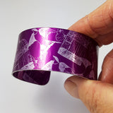 Front view of a cuff held in a hand with dark magenta background with swallows butterflies and delicate bird cages illustrations in a silvery colour. The butterflies overlap the bird cages and the birds appear to fly up from the bottom of the cuff to the top of the cuff. A very small portion of the patterned inside of the cuff can also be viewed on this photograph.