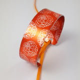 Photograph of a bright orange cuff with marigold flower illustrations in a silvery colour reapeted on the inside and outside of the cuff. The ends of the cuff  have holes in each end centrally placed and an orange ribbon through the holes and tied in a bow 