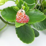 Close up shot of an orange heart shaped pendant covered with stylised silvery linear roses motif displayed on a bright green leaf.