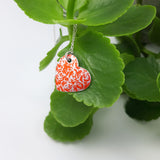 Orange heart shaped pendant covered with stylised silvery linear roses motif displayed on a bright green leaf.