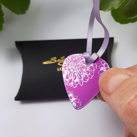 Pink anodised aluminium guitar pick pendant with silvery coloured linear drawings of aster flowers held between a finger and thumb over a black pillow shaped gift box with the Sally Lees logo in gold colour.