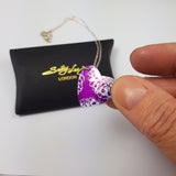 Small, pink, heart shaped, anodised aluminium pendant with portions of aster flower prints in a silvery colour displayed being held between a finger and thumb above a black pillow shaped gift box with a gold coloured 'Sally Lees' logo on the box. Photograph also shows part of the silver chain trailing from the pendant.