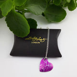 Pink heart shaped pendant with a linear print of sweet pea flowers on it with a silver chain.  The pendant is placed on a black pillow shaped gift box with the Sally Lees logo printed on the gift box in gold. In the back ground is a lucious green plant.