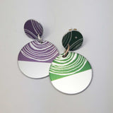 Sufragette drop stud earrings. Earrings are a small disk witj  a large disk connected by a silver jump ring. The pattern is on the top of the small disk and covers half of the larger lower disk. One earring is purple with a silvery colour and green with a silvery  colour. The patterns on each of the portions of colour are half concentric circles in a silver coloured linear pattern.