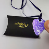 Front of a purple Guitar pick pendant decorated with a delicate linear print in a silvery colour of larkspur flowers. The pendant has a purple satin ribbon and is held in a human finger and thumb above a black pillow shaped box with the Sally Lees (London) logo in gold printed onto the box.