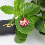 Red heart pendant with silvery coloured inear roses pattern on a green leave plant and logo box in the back ground.