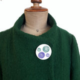 Womens suffrage round white, disk brooch with one large and one small purple disk and one small and one large green disk riveted to the main circular brooch displayed on a green coat