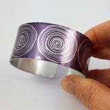 Anodized aluminum digitally printed cuff with large motifs of the end of scrolls in a linear pattern of concentric circles in a silvery colour on a purple background. The inside of the cuff is a silver colour.