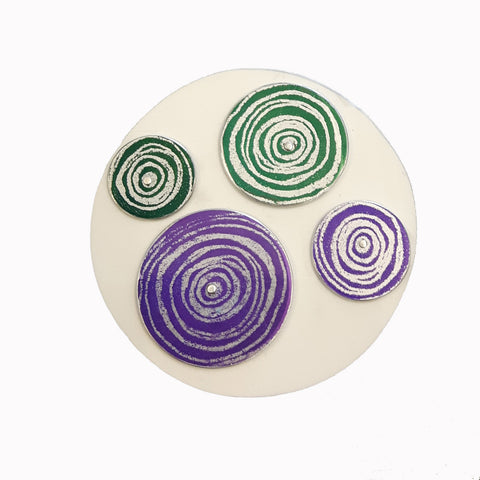 Round white aluminium brooch decorated with two purple and two green disks of different sizes with silvery coloured scroll motifs on them. The disks are riveted onto the white disk with siver rivets.