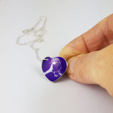 Purple heart shaped pendant with a line drawing of a cute finch seen from the side printed in a silvery colour covering the surface of the heart. The bird has a berry in its beak. The pendant is held between the index finger and thumb of a hand and the silver chain is also on display.