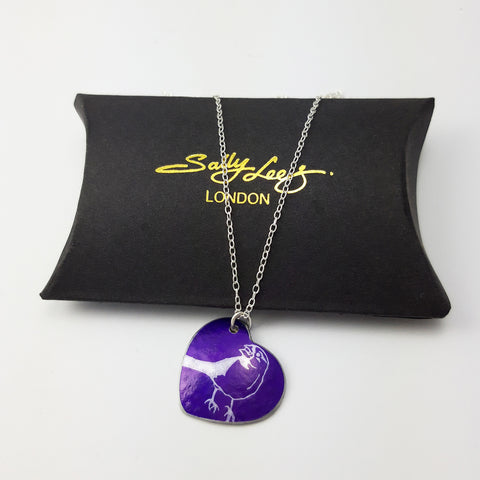 Purple heart shaped pendant with a line drawing of a cute finch seen from the side printed in a silvery colour covering the surface of the heart. The bird has a berry in its beak. The pendant is displayed on a black pillow shaped box with the Sally Lees logo embossed in gold.  The silver chain is draped over the box.