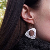 Close up of a single triangular shaped drop earring on a long silver hook wire on the ear of a model with long brown hair. The earring is patterned with a series of equally sized pattern of concentric circles.