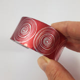 Red cuff with silvery coloured smotifs of the end of scrolls line drawings that fill the width of the cuff held in a hand.