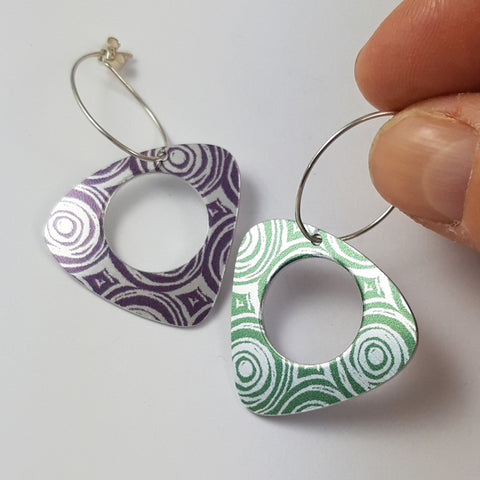 Triangular earrings with rounded edges with circular holes centrally placed in each earring. The earrings are made from anodised aluminium and are different colours - one green and one purple. The patterns on each earring are many concentric circles next to each other in a uniform pattern. The earring wires are eliptical shaped and the earring on the right is held between a finger and thumb of a human hand.
