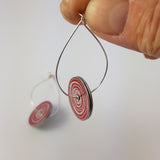 A side view of the women's suffrage earrings showing most of the red side of the disks. The silver earrings are held in between a finger and thumb at the top.