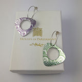 Anodized aluminum triangular earrings with rounded edges and large circular holes in the middle. The pattern on the earrings is of the end of scrolls in green on one earring and purple on the other. Each earring has a long eliptical silver ear wire. This pair of earrings is displayed on a white gift box with the Houses of Parliament logo above the Sally Lees logo all in gold.