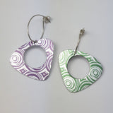 Pair of triangular drop anodised aluminium patterned with concentric circles. One earring is green with a silvery coloured pattern and one is purple with a silvery pattern. Each earring has a lage circular hole in the middle.