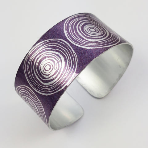 Anodized aluminum digitally printed cuff with large motifs of the end of scrolls in a linear pattern of concentric circles in a silvery colour on a purple background. The inside of the cuff is a silver colour.